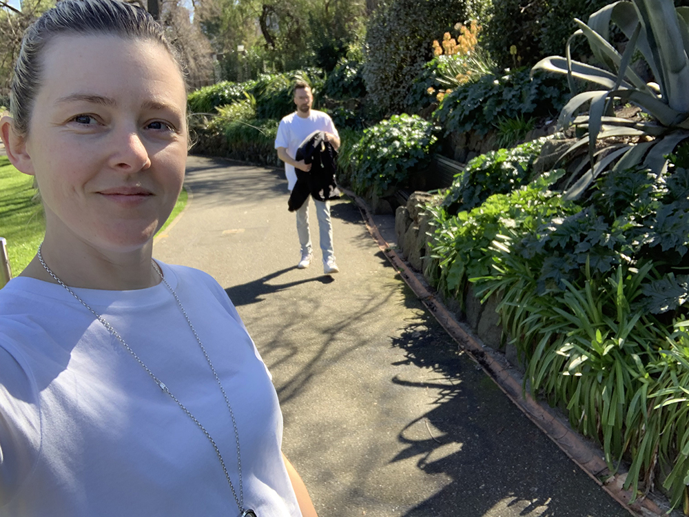 A photo of a girl wearing a white t-shirt taking a selfie in a park, with a man walking towards the camera in the background