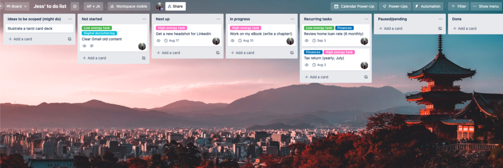 Screenshot of a Trello board, showing a photo of a Japanese city with mountains in the backdrop, behind tasks in lists (ideas to be scoped, not started, next up, in progress, recurring tasks, paused/pending, and done)
