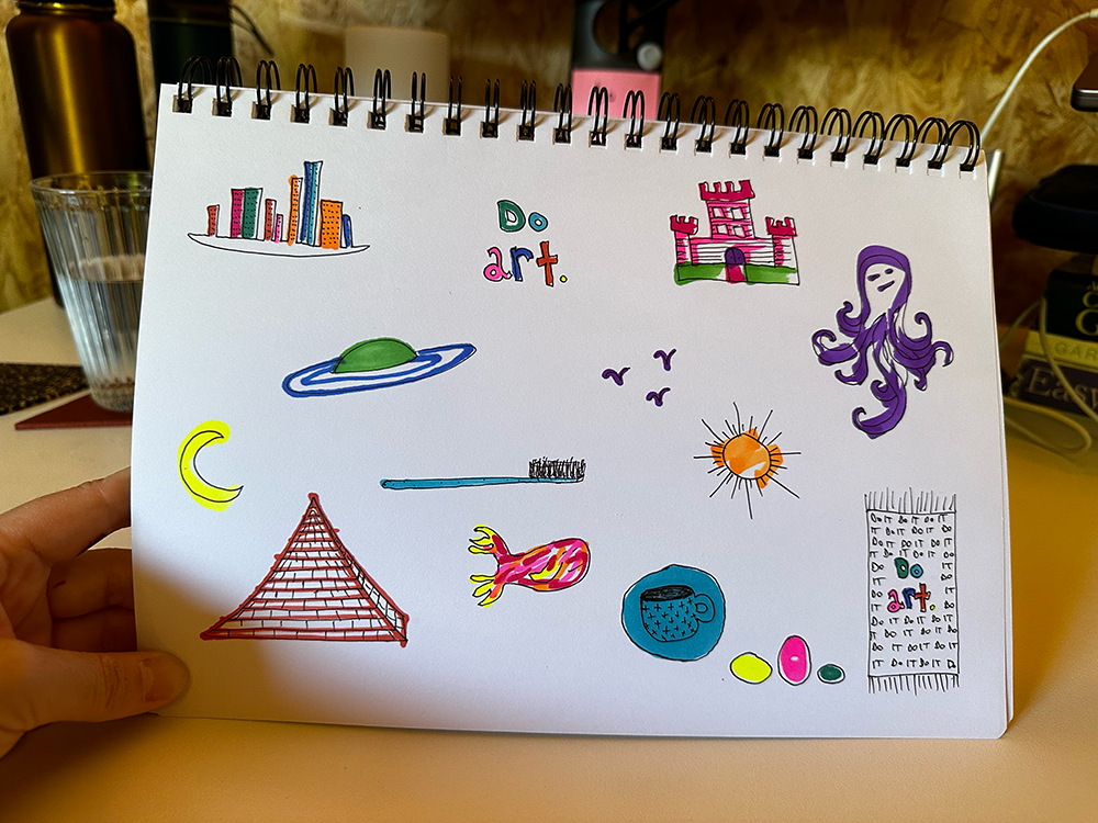 Photo of a sketchpad with an array of black line illustrations coloured in various neons, including a pyramid, an octopus and a toothbrush